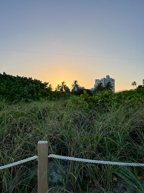 Close-up image of sand along a seashore and multi-hued sunset sky in the distance; image of an açaí breakfast bowl, journal, a framed painting, a bamboo plant, decorative wooden fruit, a vase, and a coaster on a kitchen dining table; image of tall beach grasslands in the foreground with buildings in the background backlit by the sunset light; dappled light through leaves of a mango tree and strung Tibetan prayer flags from the second-floor entrance to a yoga studio; image of a salad from a patio table and dining area surrounded by lush vegetation; image of two feet in sneakers on the sand with an infinity symbol drawn in the sand.