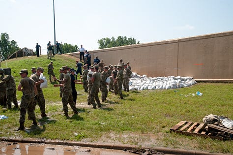 The progression of a sand boil from occurrence to remediation: 1) A soldier in fatigues carries a sandbag downhill alongside a floodway structure (a tall wall) with a cigarette between their lips and eyes turned down toward the target in the grass. The sand boil looks like a gurgling fountain a few inches in diameter, with muddy water spreading around it. 2) The floodway structure, chain of soldiers and ring dyke (several dozen sandbags stacked around the sand boil in a half-moon formation at the base of the wall) are seen from just uphill. A person in a neon yellow safety vest stands in muddy, ankle-deep water as it rises inside the ring dyke, which is now a few sandbags high. Inside the floodway structure, floodwaters as sludgy as brownie mix appear to loom several feet over the heads of the soldiers working on the landside. Two military SUVs pass on the road in the background. 3) Soldiers in fatigues pass sandbags down from pallets on a flatbed. In the foreground, a blond soldier with a square jaw and solemn expression looks on in a camouflage cap. 4) Soldiers in fatigues stand perpendicular to the floodway structure, passing sandbags toward the ring dyke. 5) Two people stand side by side, facing the ring dyke with their backs to the photographer. Muddy water spills over the sides of the ring dyke, which now stands five or six sandbags high. The person on the right is in fatigues. The person on the left wears a neon yellow safety vest with the red and white turreted castle emblem of the U.S. Army Corps of Engineers. 6) Several members of the Oklahoma National Guard stand in, on and around the ring dyke in fatigues. One is submerged to calf- or knee-height in the muddy water spilling over the edges of the ring dyke. Another steps calf-deep into the pool with one leg, straddling the ring dyke to position a sandbag.