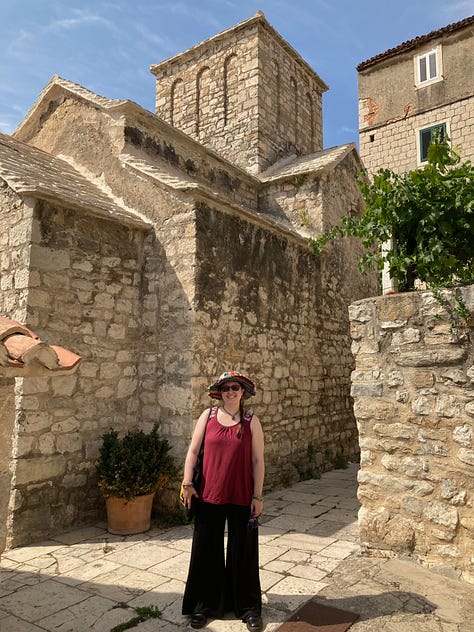 A woman in a red and black dress, bending down to look into a drainage hole in a wall in Pendragon Castle, a 12th-century castle in Cumbria, England; A woman wearing sunglasses, peering through a medieval squint at Penmon Dovecote, built c. 1600, near Beaumaris in North Wales; a woman wearing a red t-shirt, black trousers and a hat standing in a walled street in Split, Croatia. 