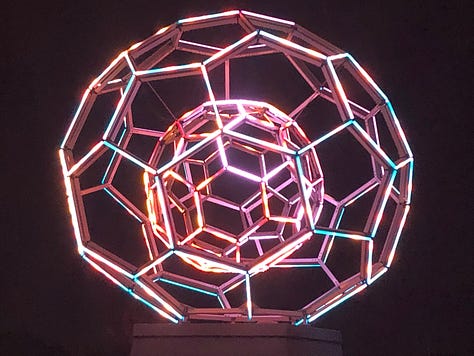 Blue faerie lights kiss the forest ground. Vertical light bars pierce into the night in 80s neon hues. An LED soccer ball within a soccer ball glows turquoise, gold and fire.