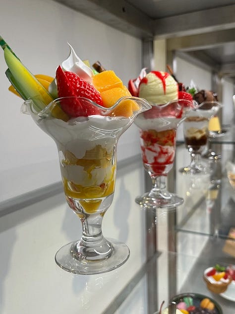 Three examples of colourful, Japanese replica dishes including a bento box and two desserts with fruit, cherries and cream