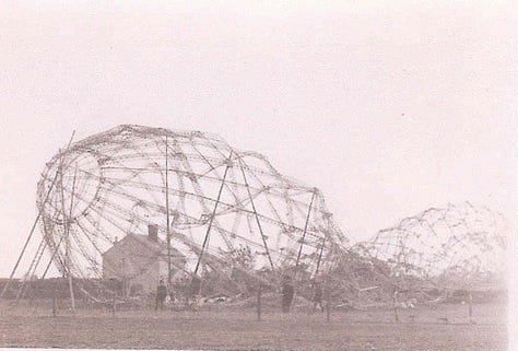 The zeppelin's metal scaffold structure lays in a field, several stories high. Officers inspect the structure and a crowd waits behind a taped cordon.