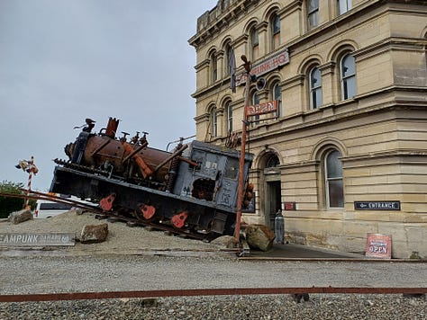 A series of stone buildings, created in a variety of styles, from fairly plain and study to huge and ornate. All of the buildings are made from or incorporate creamy limestone, some soot-blackened or weathered. On the right top is a locomotive engine cab with a large screw mounted at the front, located to indicate that it has just burst up out of the ground. The bottom right features a whitestone bridge fording a stream, over which the main street is laid.