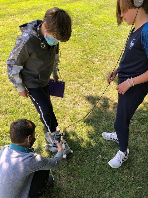 kid sitting at a desk doing math, kid lying on the floor with a laptop doing situps, three kids holding a microphone and recorder, interviewing a stuffed bear