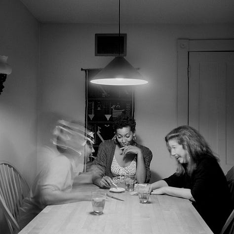 1-9) All images are black and white, and involve a table, as well as a few chairs, and an overhead light. 1) A black woman is seen playing cards by herself. 2) A black woman is seen holding her head in her hands, while reading a book. There is a child sitting to her right, writing. 3) A black person is standing at the end of the table, looking into the camera. 4) A black woman is looking at herself in a tabletop mirror, while a child is playing to the right of her at the table. 5) Three people are sitting at the table, and appear to be playing a game or laughing. 6) A black woman is seen looking at herself in a tabletop mirror. 7) A black woman is sitting on the left side of the table, with her hand under her chin. There is a black man at the end of the table, reading the newspaper, with his hand resting on his neck. 8) Two black people are seen kissing/embracing. One is sitting at the end of the table, and the other person is behind them. 9) A black woman is seen with her head laying back on the table, with her hands in her hair.