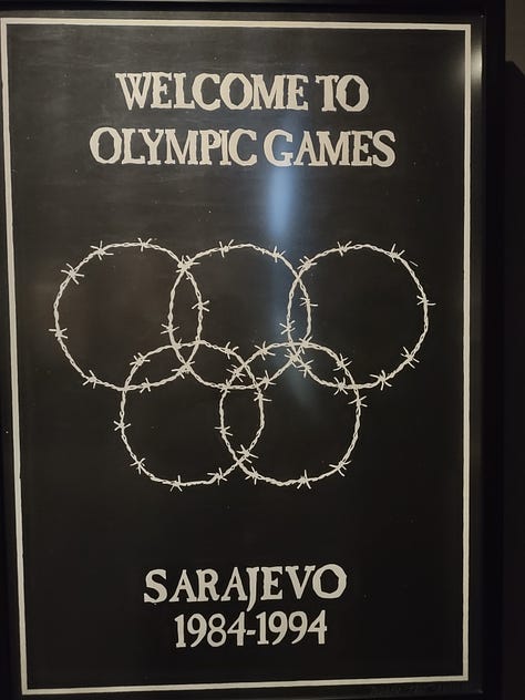 The top left image shows a black poster with barbed wire rings in the form of the olympic rings. It says "welcome to the olympic games. Welcome to Sarajevo". The top middle image shows a sign that says "enjoy Sarajevo. 1993/94" in the shape of the coca-cola logo. In the bottom it says "coca-cola logo redesigned by trio sarajevo". The top right image shows a picture of a Campbell's Soup can that says "Sarajevo';s condensed chicken with rice soup." The cna has bullet holes in it. Underneath it says "Andy Warhol Campbell Soup redeisnged by Trio Sarajevo." The bottom left image shows a poster on a blue background with the united nations logo. It says "1993. Disunited nations of Bosnia and Herzegovina." The bottom right image is of a woman on a black background with stars in her hair. She is sleeping. It says "wake up, Europe! Sarajevo calls every man woman and child."