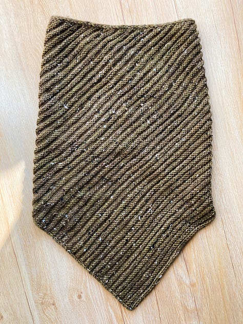 A brownish-green knitted cowl laying on a light brown floor in different positions.