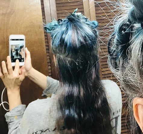 The author getting ready to re-dye and re-bleach her hair in sea-blues, pulls up the back of her hair where it's still brunette, in contrast to the silver roots on the top and sides of her head.