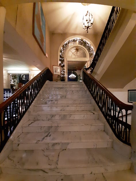 A collage of photos showing the Historic Hotel Bethlehem, including two photos of the 9-story brick exterior, and interior pictures of the elevators, white marble staircase with polished handrail, open doors to the ballroom with a glistening chandelier, and two photos of the lobby, with luxurious black leather furniture in front of a huge arched window with a Christmas wreath in the middle. Also a look inside a room of a bed with a robe lying on the end.