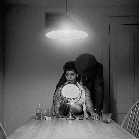 1-9) All images are black and white, and involve a table, as well as a few chairs, and an overhead light. 1) A black woman is seen playing cards by herself. 2) A black woman is seen holding her head in her hands, while reading a book. There is a child sitting to her right, writing. 3) A black person is standing at the end of the table, looking into the camera. 4) A black woman is looking at herself in a tabletop mirror, while a child is playing to the right of her at the table. 5) Three people are sitting at the table, and appear to be playing a game or laughing. 6) A black woman is seen looking at herself in a tabletop mirror. 7) A black woman is sitting on the left side of the table, with her hand under her chin. There is a black man at the end of the table, reading the newspaper, with his hand resting on his neck. 8) Two black people are seen kissing/embracing. One is sitting at the end of the table, and the other person is behind them. 9) A black woman is seen with her head laying back on the table, with her hands in her hair.