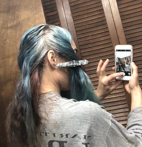 The author getting ready to re-dye and re-bleach her hair in sea-blues, pulls up the back of her hair where it's still brunette, in contrast to the silver roots on the top and sides of her head.