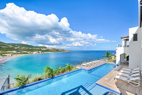 An aerial view of Villa Amalia perched on the cliffs of Oyster Pond, St. Martin, showcasing its sleek modern design with an expansive infinity pool overlooking the turquoise Caribbean Sea. The villa’s open-concept layout, floor-to-ceiling glass walls, and spacious outdoor terrace offer a perfect blend of indoor and outdoor luxury living, with lush greenery surrounding the property and the ocean visible in the distance.