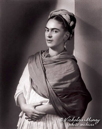 Three images of black-haired Frida Kahlo in black-and-white and color photos. Kahlo on the left in the black-and-white photo  shows her  painting. Kahlo in the middle color photo wears a purple ribbon threaded in her hair gold jewelry, and a dark pink cape. Kahlo in the right black-and-white photo leans back and wears a scarf and has a ribbon threaded in her hair.