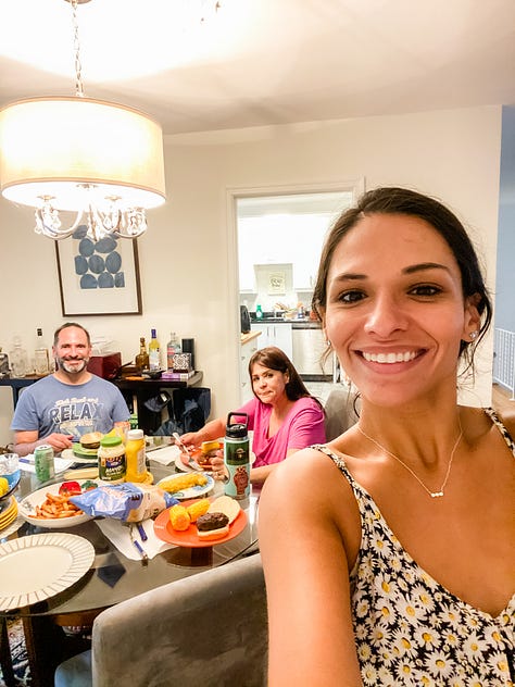 Photo of a paved walking trail surrounded by lush vegetation and a deer on one side, a round embroidery hoop with an image of a pineapple, a woman smiling and presenting the piles of fresh pasta she made from scratch, a selfie photo of woman and her dad painting a piece of furniture, a photo of a woman and her mom sitting on the floor painting a piece of furniture, a selfie photo of a woman and her parents sitting down at the dinner table to eat barbecue food, a photo of a woman and her dog looking out the window, a photo of a LOVE sign in a grassy, woody green forest, a selfie photo of a woman with her parents and dog behind her walking in the opposite direction.