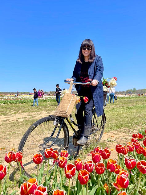 Left: Jenny in a blue sweater and dark jeans on a bike with some tulips. Center: Jenny in an all-black getup with sunglasses, a mask, and tote bag. Right: Jenny sitting outside in some cherry blossom pants and blue-gray sweater.