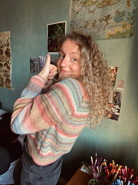 JB wearing a pastel coloured striped jumper that she knitted. She is standing in said jumper looking very pleased, with her thumbs up and a smile on her face.
