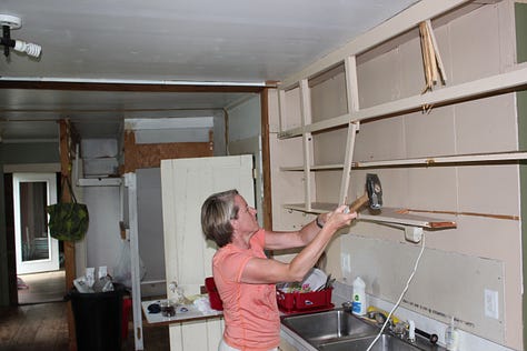 woman wields a sledge hammer to shelves, a new refrigerator stands in a room gutted to the studs, a farmhouse kitchen with a red woodstove, an old beam, a Yellow Lab and wood floors