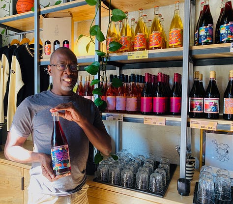 Wine bottles with images of my artwork on the labels arranged in rows on shelves. In the middle is a photo of me, a man holding a bottle of the Pet-Nat Rouge smiling for the photo and looking out at the viewer.