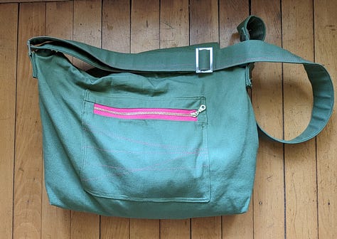 Five photos of the Mythical Messenger Bag. In the first, I (a white lady, aged 52) am standing in the hallway wearing the bag. The other photos are of the bag on its own. The bag is olive green canvas with an adjustable strap. There is hot pink decorative stitching. There is an exterior welt pocket on the back with a hot pink zipper. There is a pocket on the front with a blue square patch that says "Make Art" in a red cursive font. The inner lining fabric is floral -- blue, green, coral line drawn flowers on a white background. Inside the bag, there are two pockets. One is a zippered pocket with a hot pink zipper. The other is a divided pocket with slots for pens, a phone, and something else roughly cell-phone-sized.
