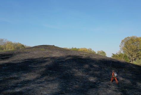 Fire burns in an open grass field, photo of fire and smoke from above, a person in orange jumpsuit walks up a blackened hill