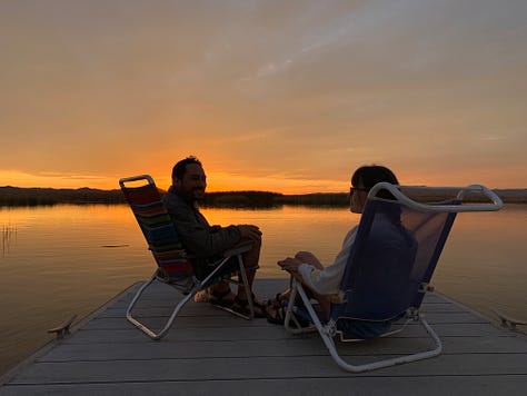 A man and a woman sit in beach chairs on a dock before a sunset. A series of sunset shots from yellow to marmalade to pink and purple to blue to fiery orange.