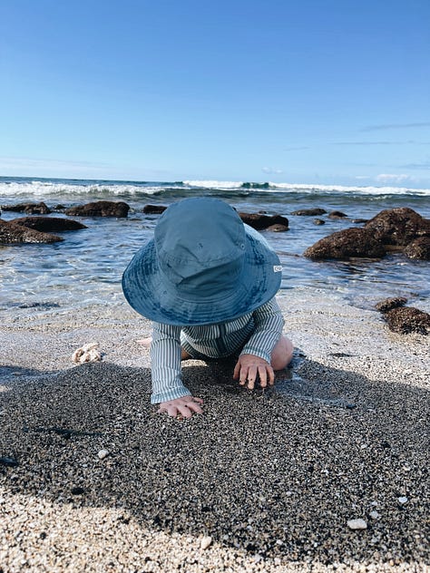 a toddler in a blue and white striped bathing suit and a blue hat plays in the sand on a beach. 