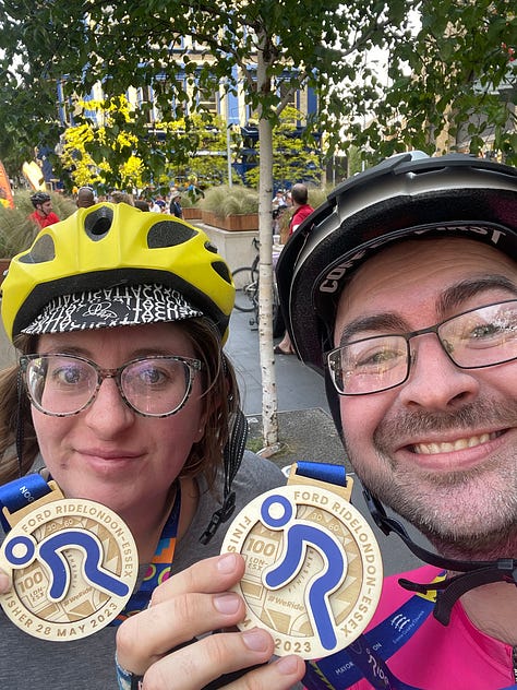 Jem and Jack smile to the camera holding medals from the cycle event. They start off in a line of cyclists on a leafy green street.