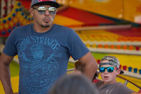 Nine photos from a fair. A closeup of the hand of a quilter; a man and a boy with blue sunglasses in front of a ride; a smiling face; human feet walking behind horses with dust; horse feet; men pulling hard on their horeseteam; horse team leaping out to pull weighted sled. two oxen standing in the sun; a young girl trying to wrestle one of the oxen on her team