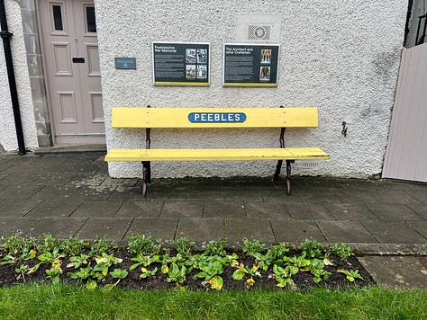 a bright yellow wooden bench with Peebles on a metal sign sits in the Quadrange off Peebles high street near the war memorial.