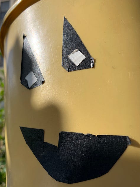 Mustard-yellow small cylinder bucket with wire handle, jack-o-lantern face adhered in black tape