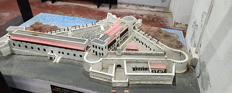 A model of Cape Coast Castle with white stone walls, and red roofing, An outdoor image on the castle grounds showing white stone and concrete pavement. An inscription in the castle wall entitled "In Everlasting Memory."