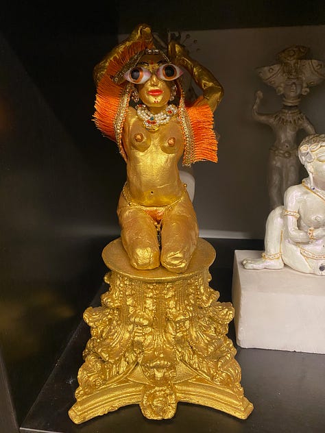 Blue goddess statue, working class woman and her daughter both in pink hijabis, gold-colored goddess icon with orange detail.