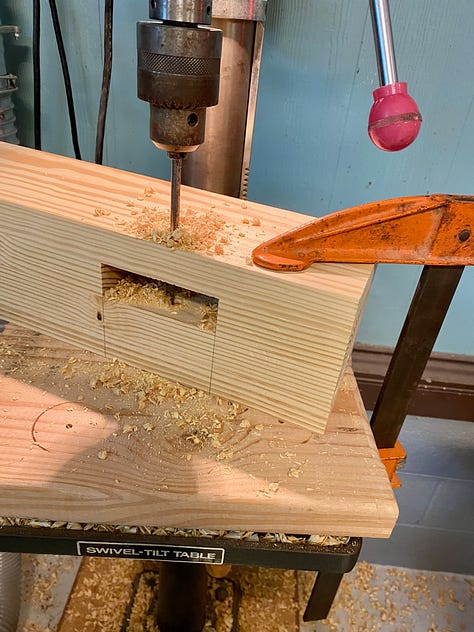 Holes bring drilled, via a drill press, into the bench legs; overlapping drawbore holes; sanding drawbore pegs; and then waxing and temporarily inserting them into the bench base.