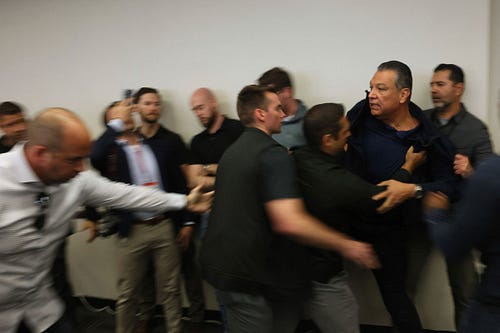 A group of people push a lawmaker out of a room during a press conference. People standing near a wall watch and take video on their phones of the incident.