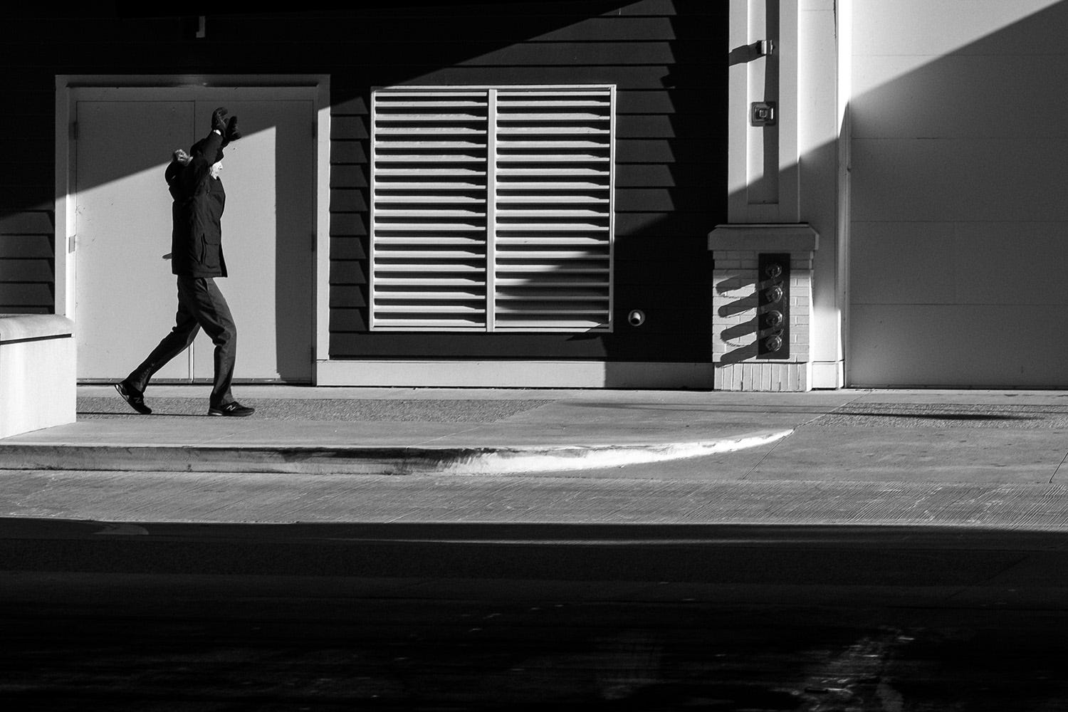 A man is walking down a light and shadow filled sidewalk. He is reaching over head similar to warm up exercises. 