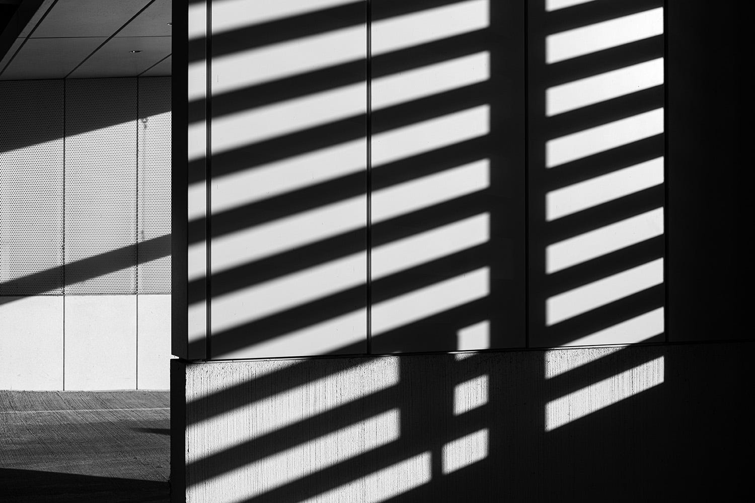Light pours through the slatted windows of a parking garage casting bold striped shadows throughout the scene. 