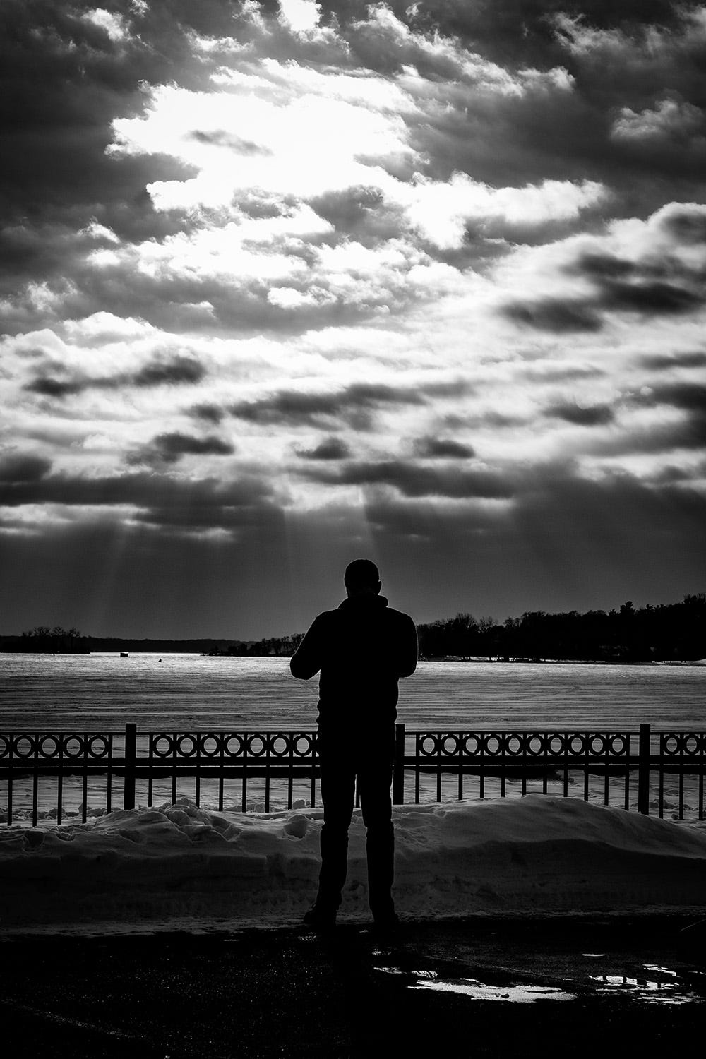 A man stands at the lakefront observing sun rays leaking through moody overcast skies. 