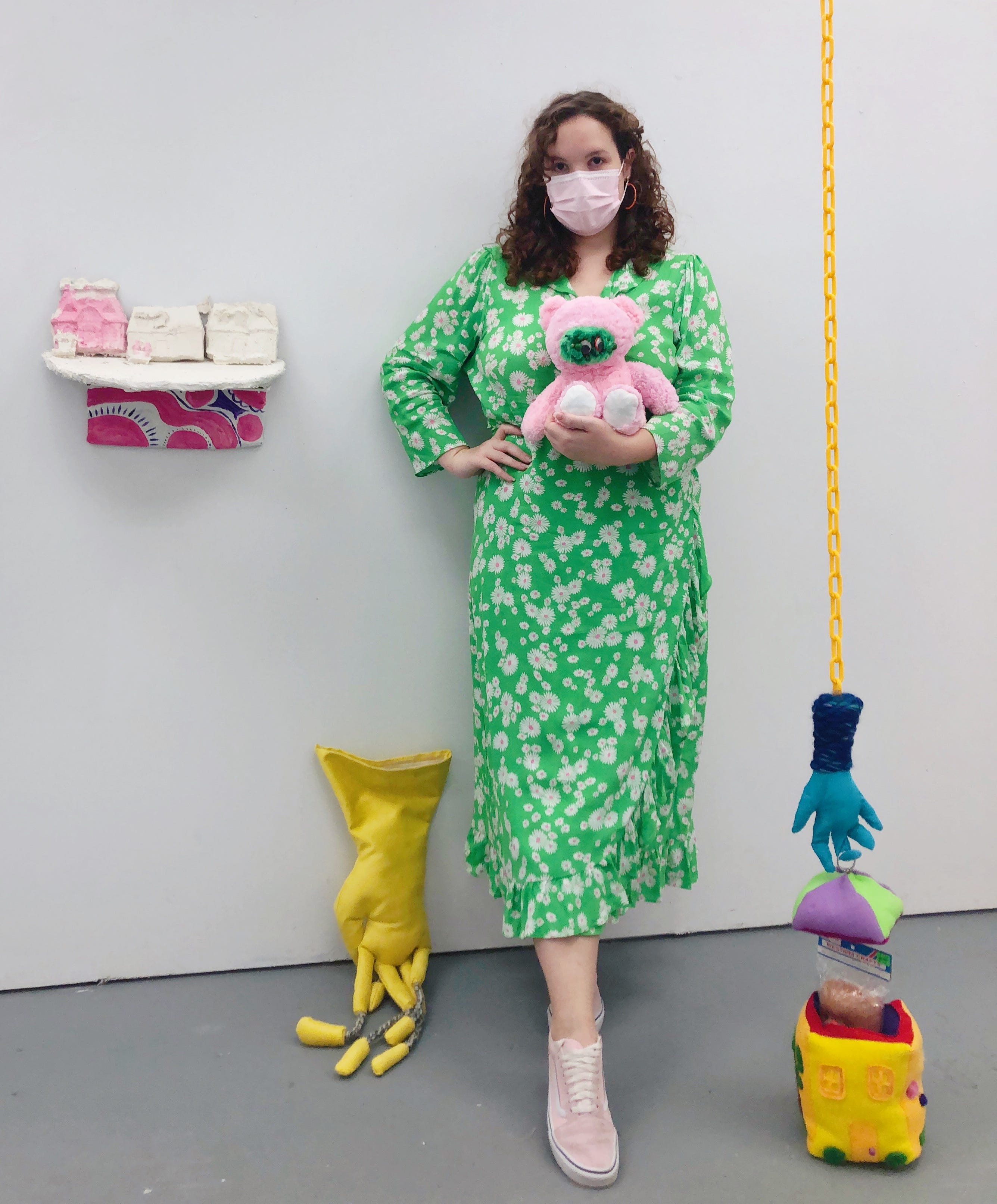 I stand wearing a mask and holding the bear which has a green face while wearing a matching green dress. I am surrounded by my sculptures which depict homes and a hand