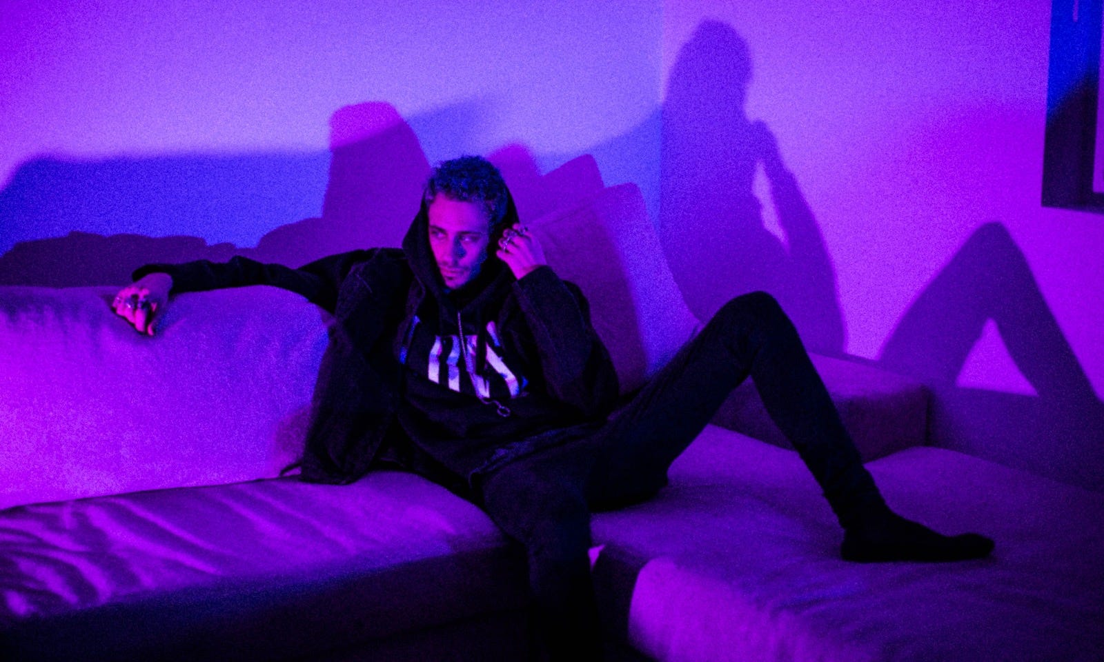 cøzybøy dressed in all black sitting on a sofa in front of a moody purple and blue background.