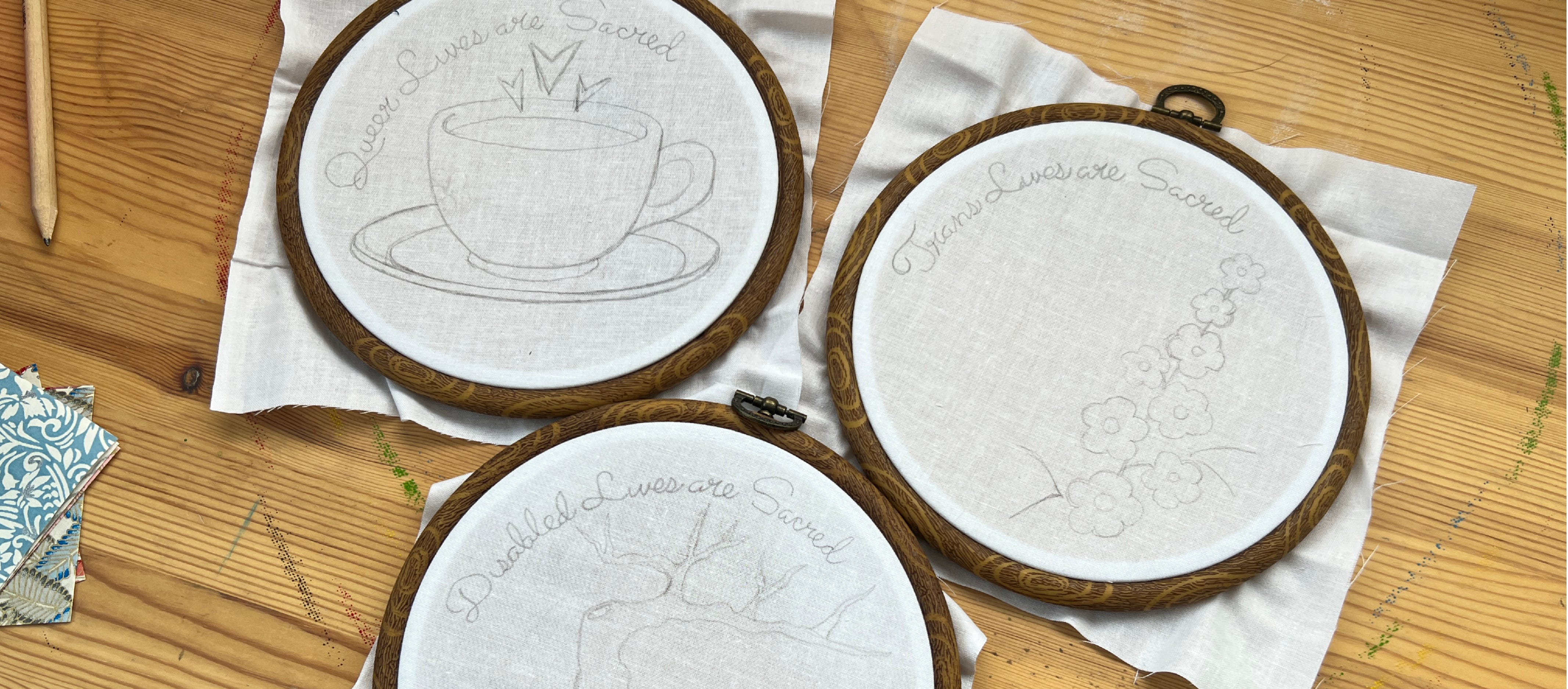 Photo of three embroiery hoops with white cloth in them and pencil sketched designs sitting on a wooden desk. The thee designs each have text, reading: Queer Lives are Sacred, Disabled Lives are Sacred and Trans Lives are Sacred. 