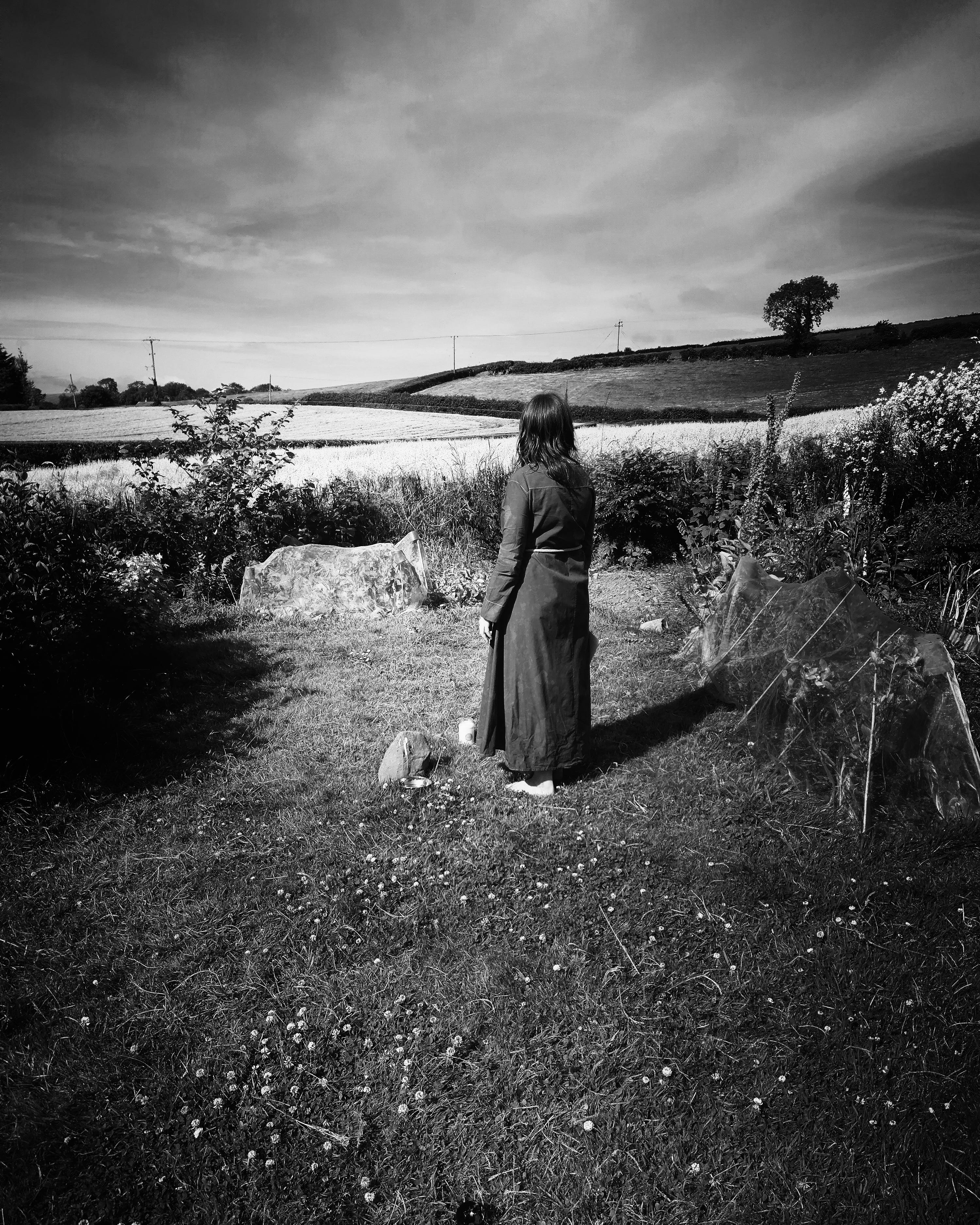 A wide black and white photo of a woman in la long cotton robe standing in the middle of a garden in front of a small standing stone. Surrounding the garden is a vast expanse of sky and undulating fields and hedges. A wide black and white photo of a woman in la long cotton robe standing in the middle of a garden in front of a small standing stone. Surrounding the garden is a vast expanse of sky and undulating fields and hedges.