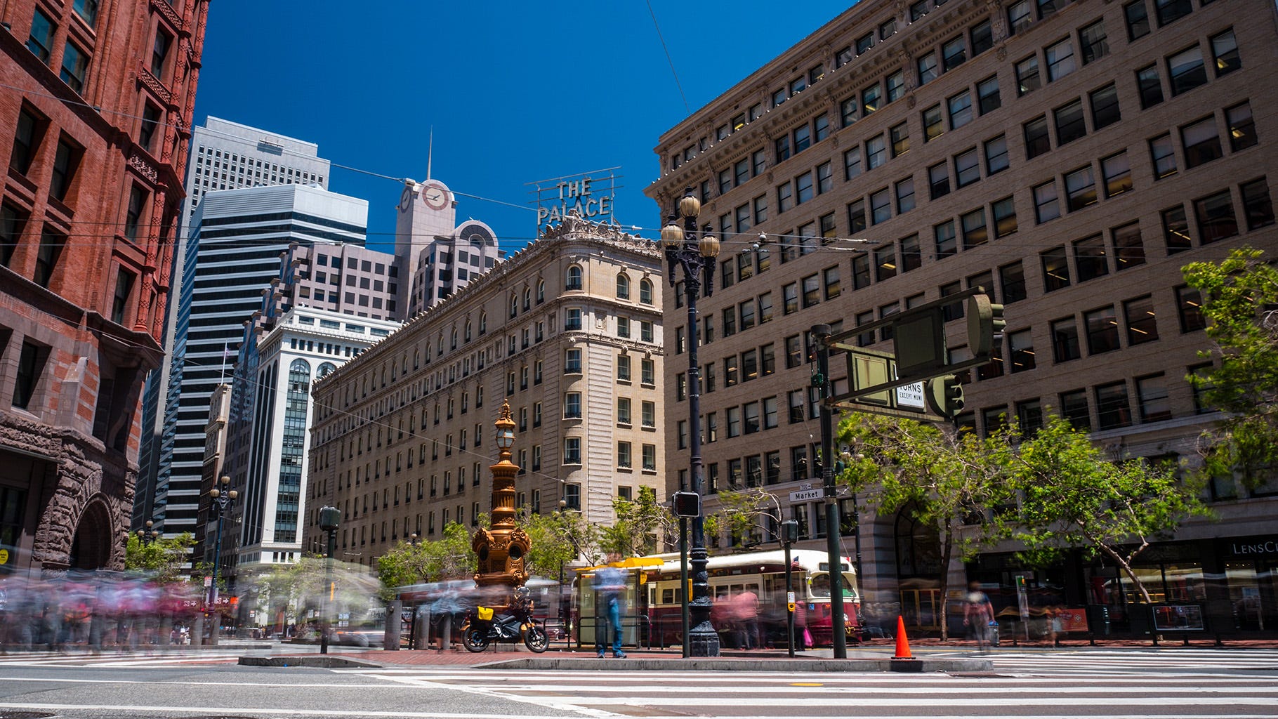 Wide-angle photo of Lotta's Fountain in San Francisco, with tall buildings surrounding a small triangular island where the bronze-colored fountain stands. In front of the fountain, a Zero electric motorcycle is parked. And on sidewalks around the street, the blurred images of pedestrians are visible (this is a long exposure photo that renders movement as blurs)