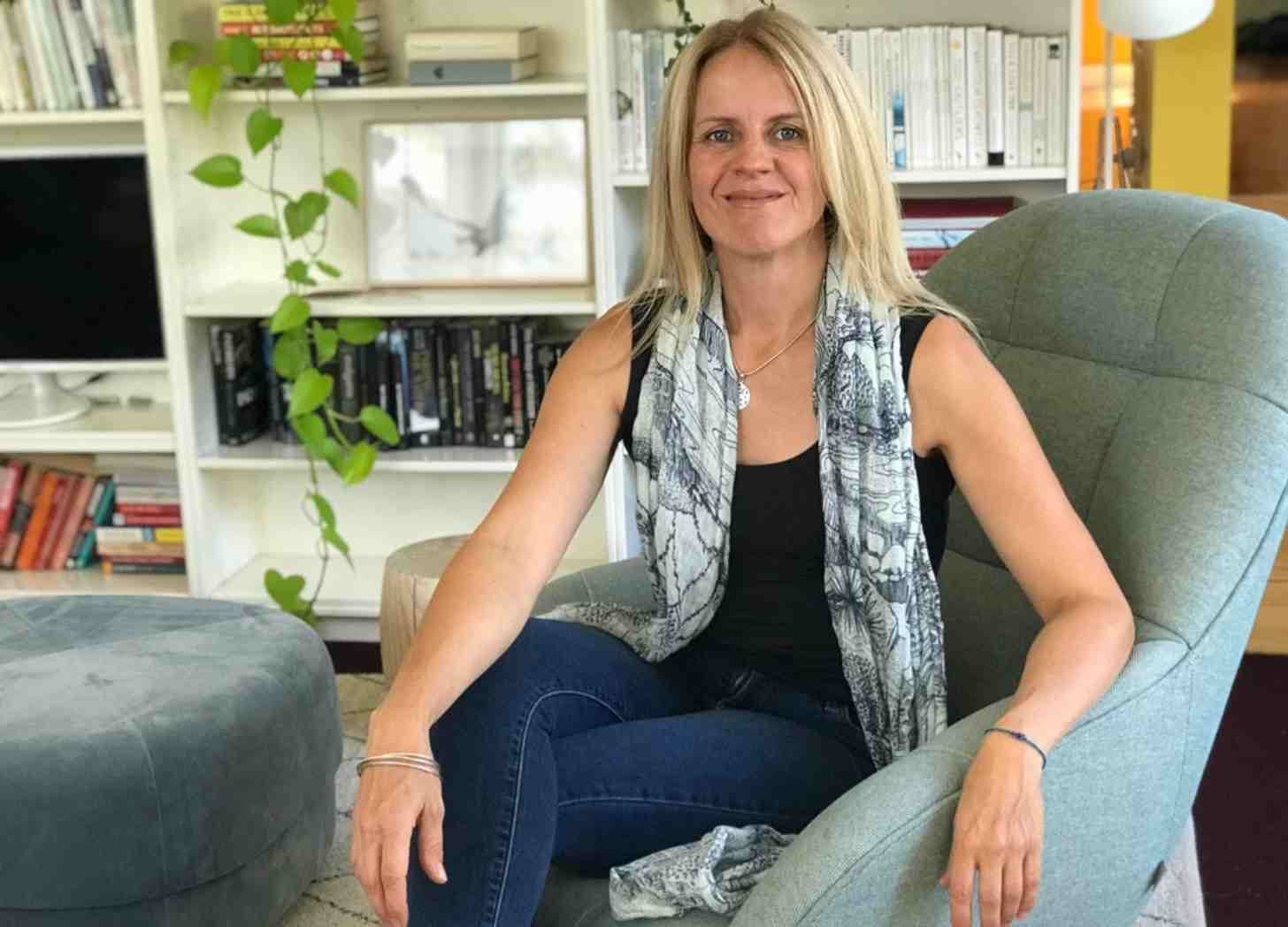 A smiling woman sits on a green armchair in front of a bookcase