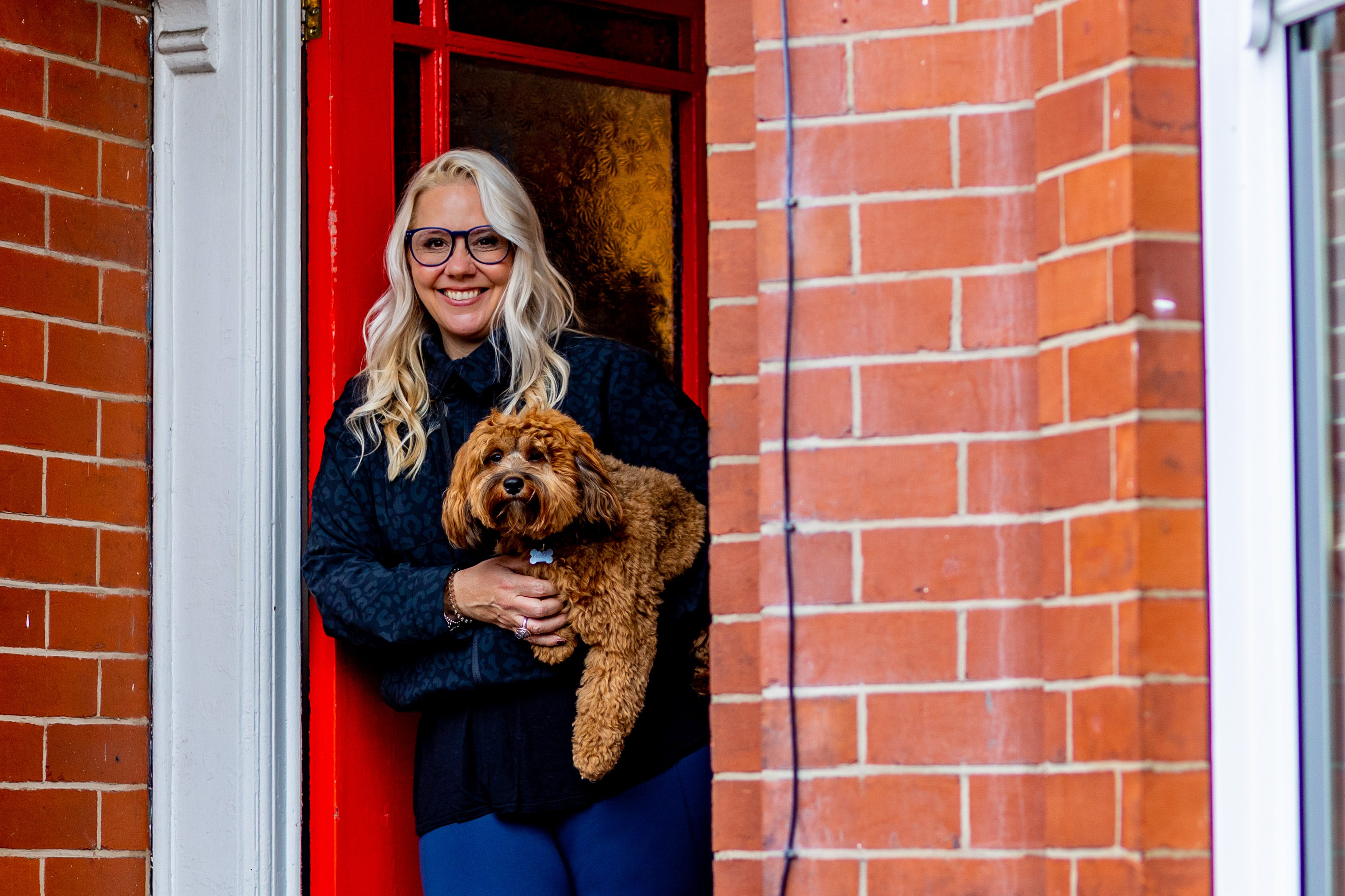 Sarah Knight is standing against a red front door, holding a gorgeous fluffy doddle-like dog. 