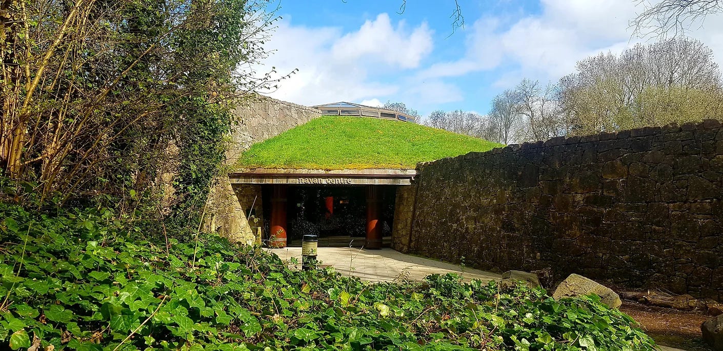 Navan Fort Visitor Centre, built underground in a grass covered mound like structure, tall wall to right of picture, ivy trees and shrubs to left.