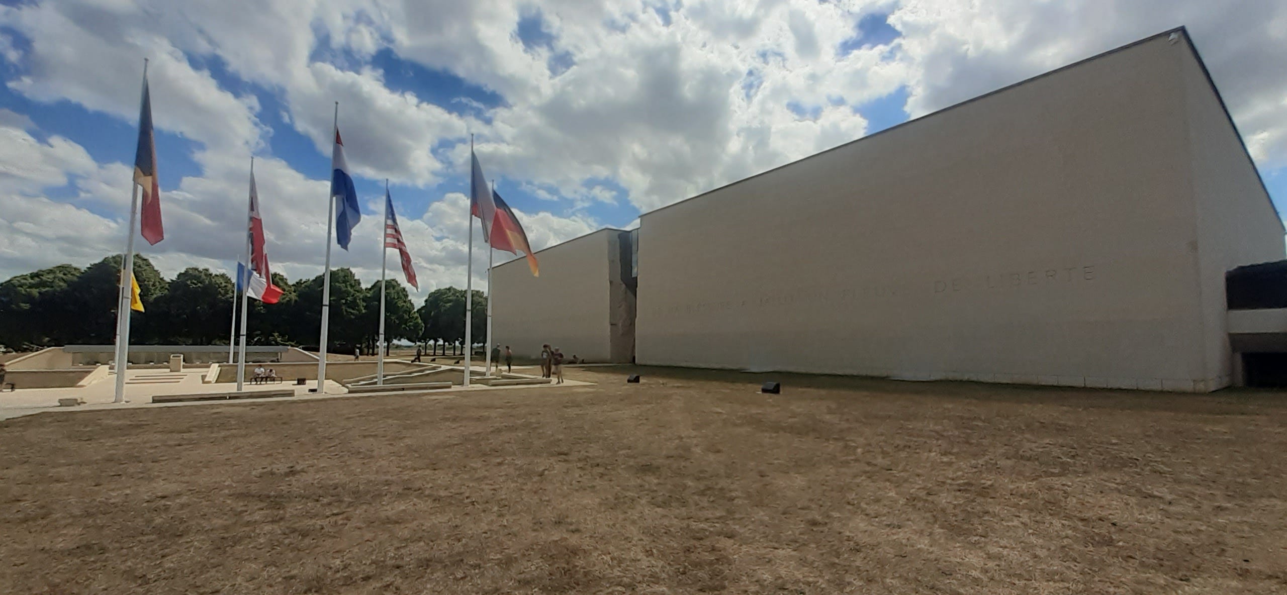 La facciata frontale del Memoriale di Caen: un parco un po' secco per la calura estiva e diverse bandiere internazionali che sventolano all'entrata. Le mura sono grigio chiaro e lisce, come un unico blocco di cemento.