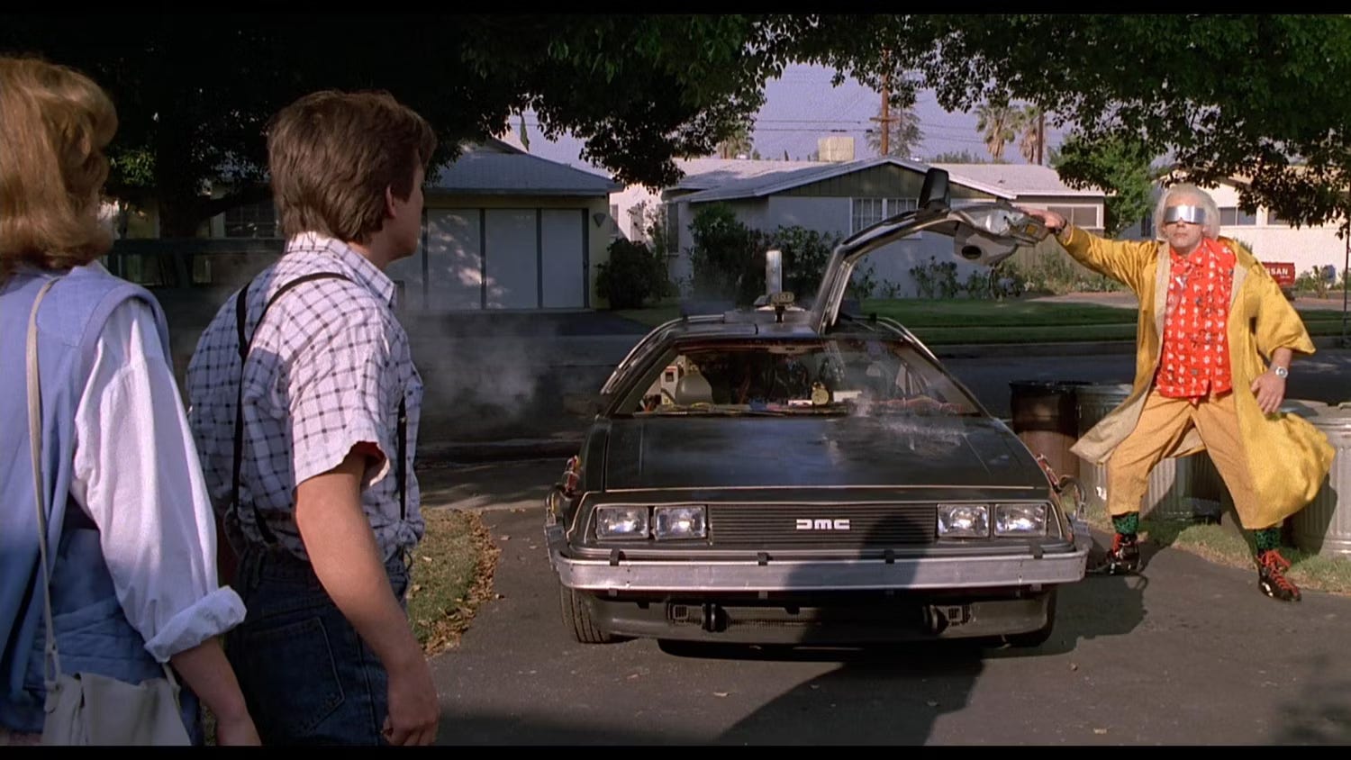 Screenshot from Back to the Future. Doc is standing to screen right, wearing a yellow suit and silver glasses, with his right hand resting on the open door of the Delorean. Marty and Jennifer are shown from the back on screen left, looking at Doc.