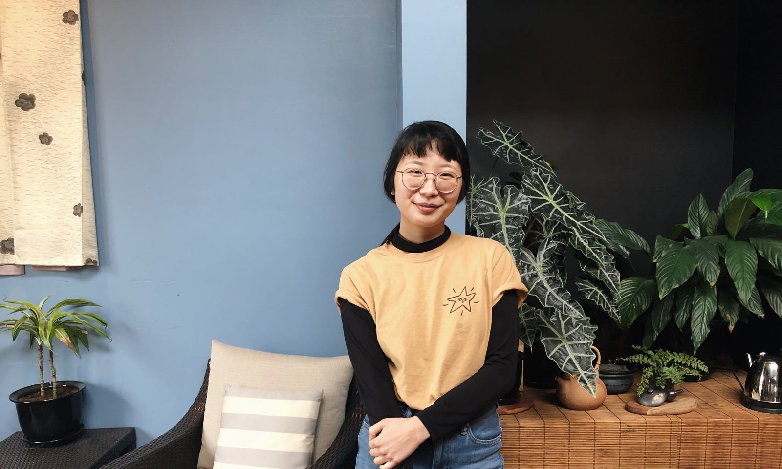 Tiffany Trieu in yellow shirt posing in front of blue wall and indoor plants.
