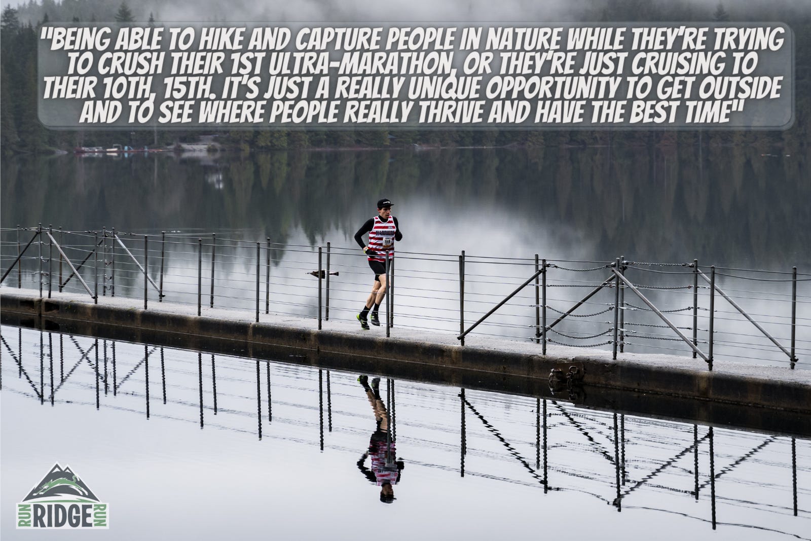 A runner crosses a narrow bridge with a perfect reflection of themselves on the water. The day is moody with clouds and grey