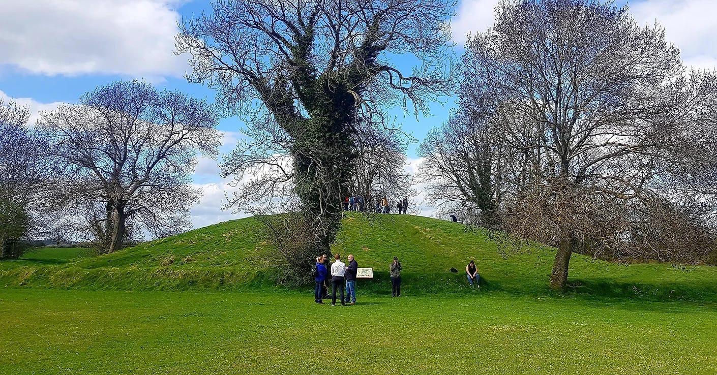 The huge mound at Emain Macha, with some people standing chatting in the foreground, surrounded by trees below a blue sky